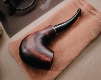 Puede incluir: Una pipa de madera marrón con una boquilla negra que descansa sobre una bolsa de gamuza marrón. La pipa tiene una cazoleta curva y un acabado liso.