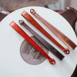 May include: Three leather camera straps in different colors: red, brown, and black. Each strap has the word "Camera" embossed on it.