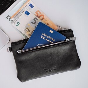 May include: A black leather wallet with a zipper closure. The wallet contains a passport, cash, and a credit card. The passport is blue and has the word "UKRAINE" printed on it. The cash is in Euros.