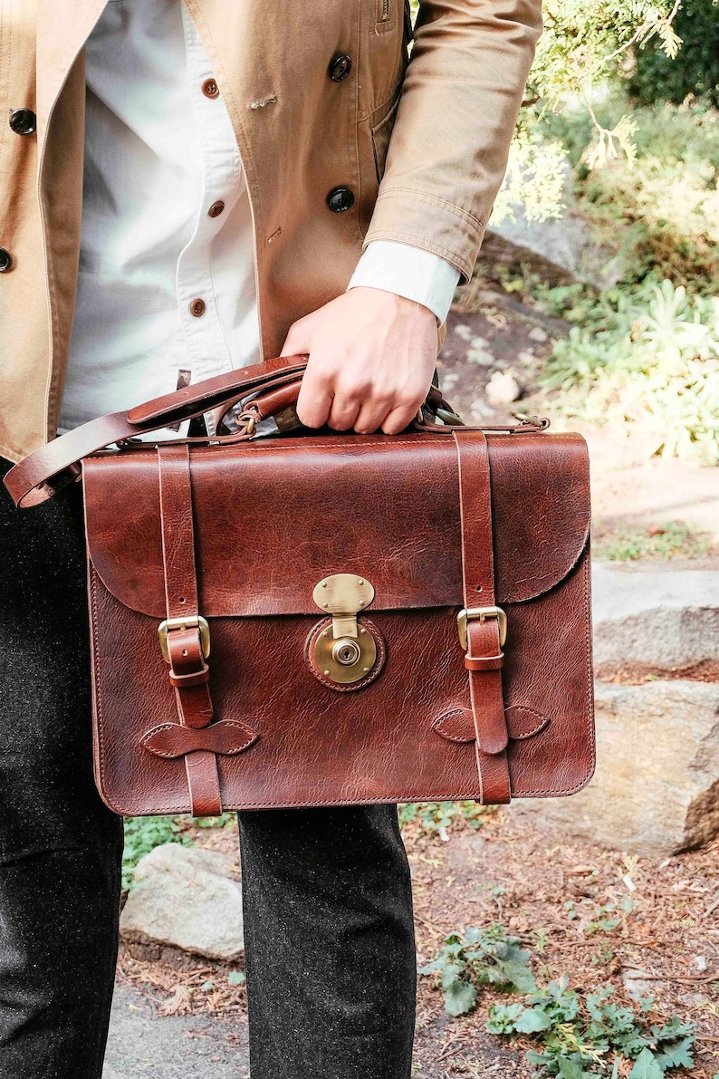 May include: A brown leather briefcase with a brass lock and buckles. The briefcase has a long strap and is being held by a person.