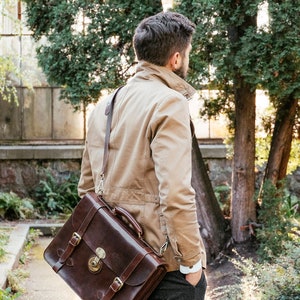 May include: A man wearing a tan jacket and gray pants carries a brown leather briefcase with a long strap over his shoulder.