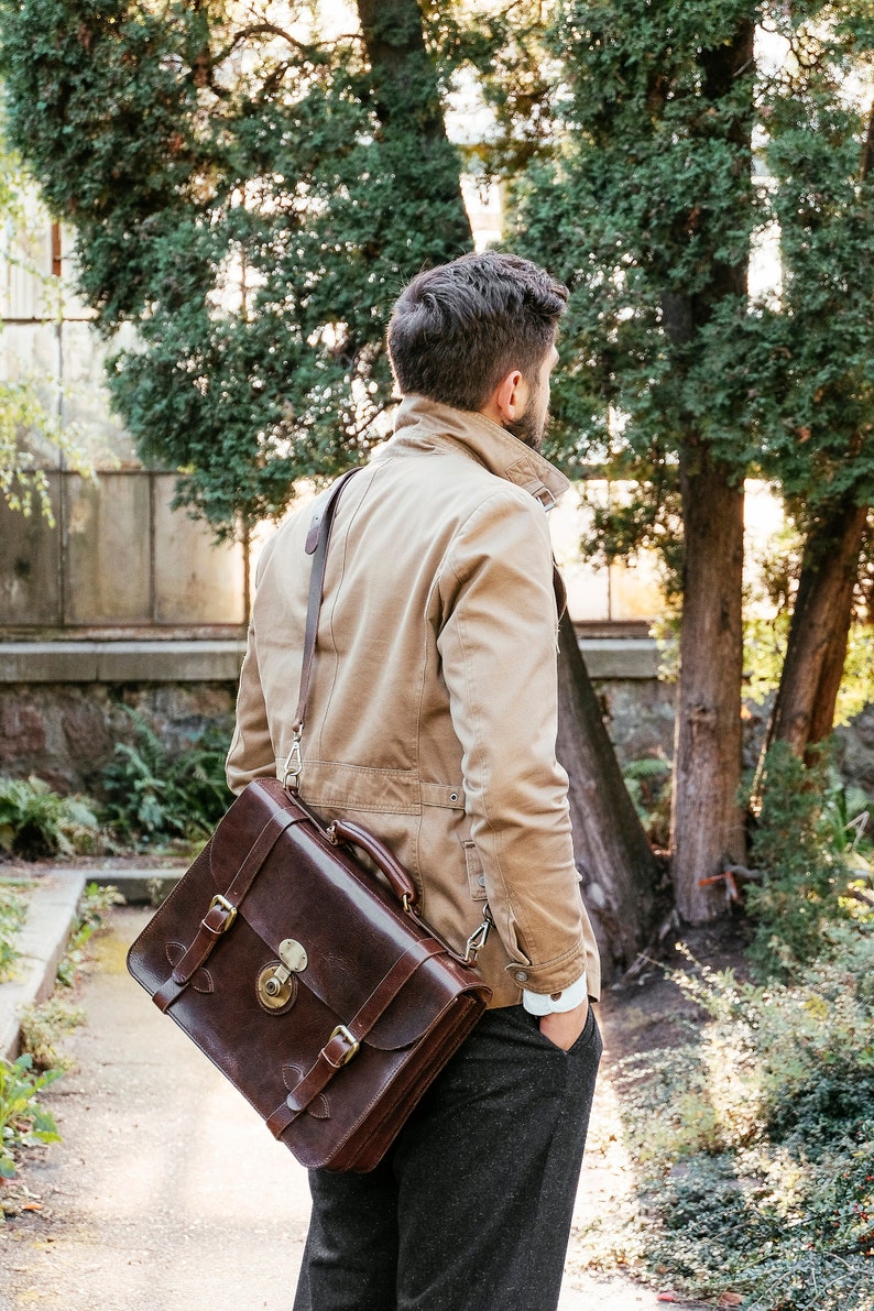 May include: A man wearing a tan jacket and gray pants carries a brown leather briefcase with a long strap over his shoulder.