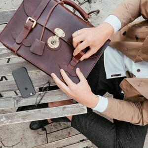 May include: A brown leather briefcase with a brass lock and buckle closures. The briefcase is open and a person is holding it.