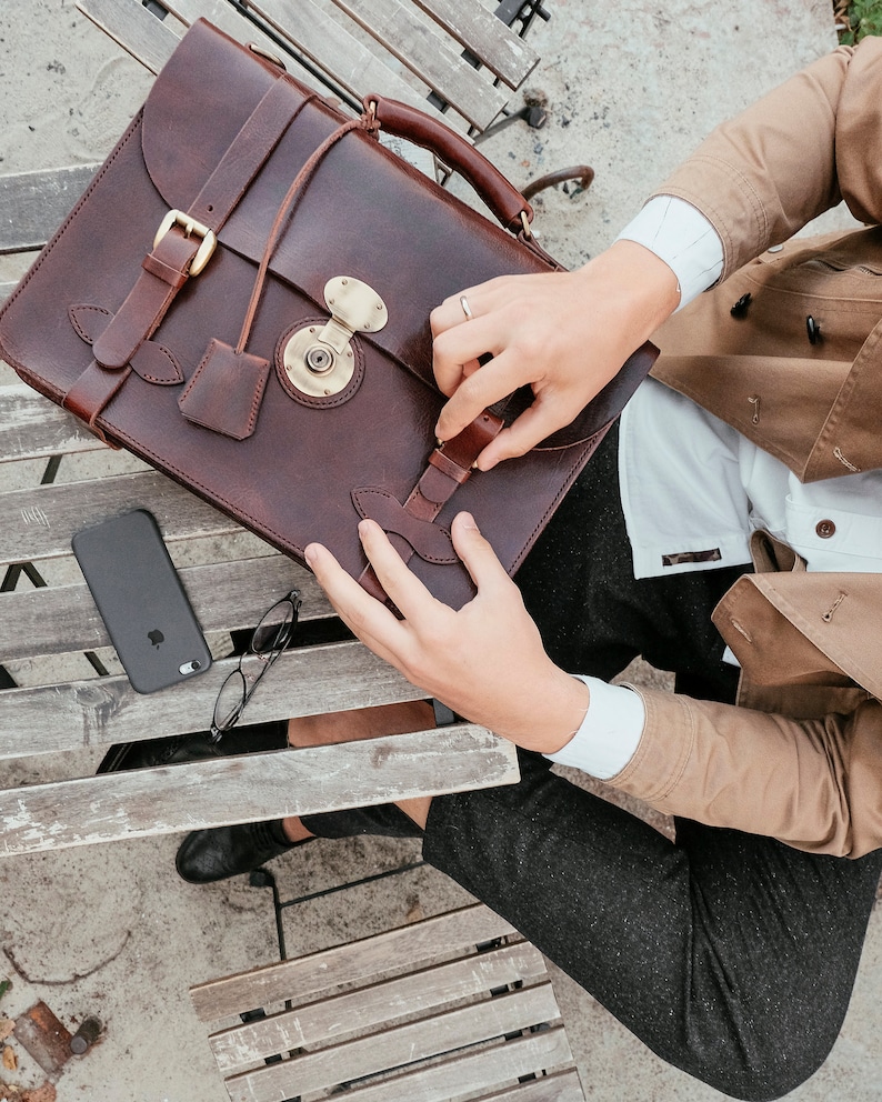 May include: A brown leather briefcase with a brass lock and buckle closures. The briefcase is open and a person is holding it.