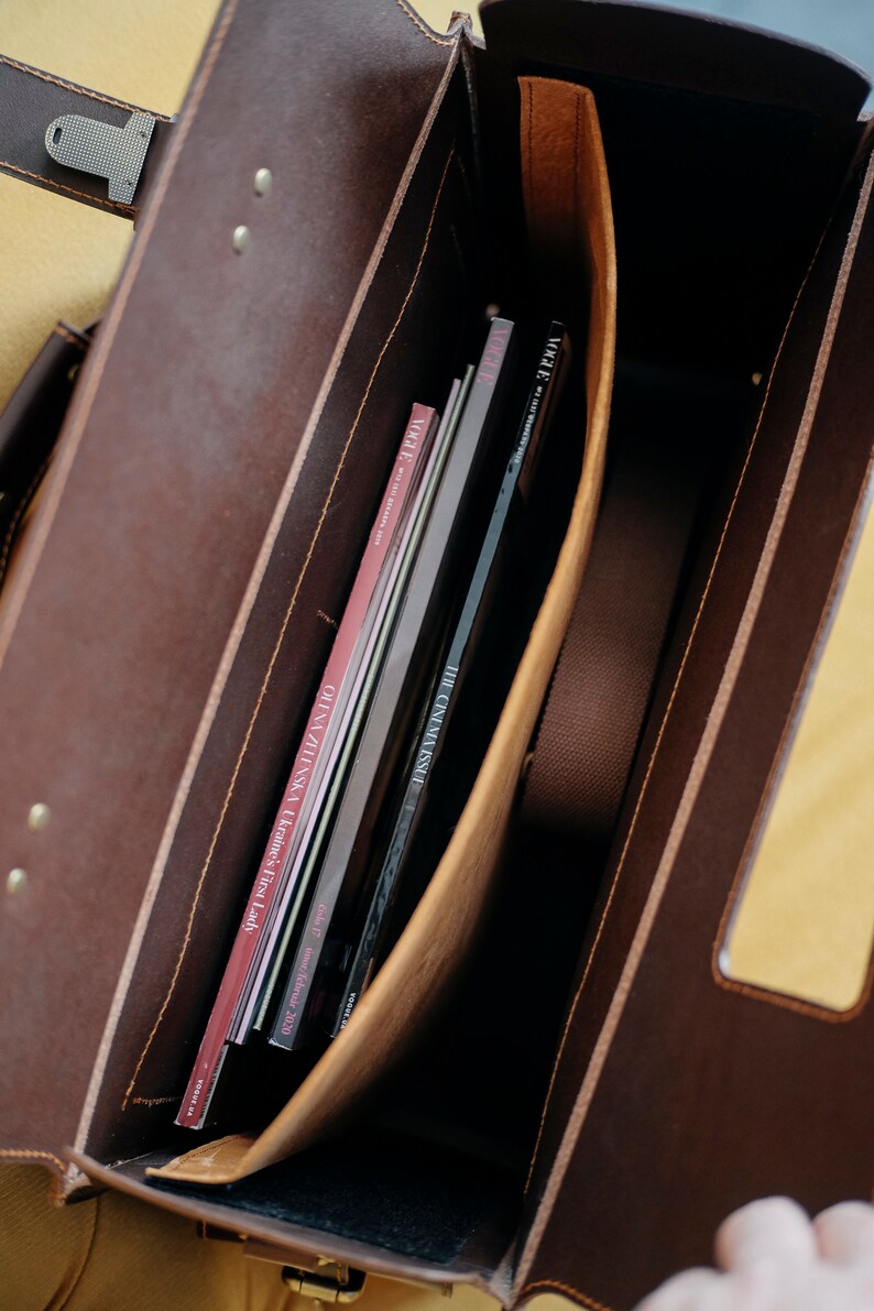 May include: A brown leather briefcase with a flap closure and a brass buckle. The briefcase is open and contains several magazines and books, including "The Cinema Issue" and "Olga Zilinska Ukraine's First Lady".