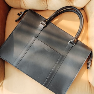 May include: A black leather briefcase with a top handle and a front pocket. The briefcase has a zipper closure and a small metal tag with the word "Handmade" on it.