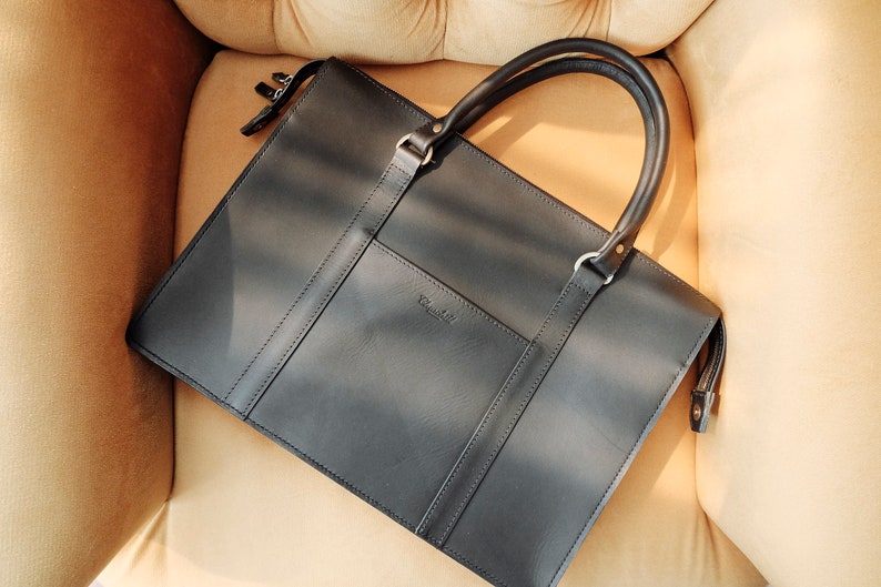 May include: A black leather briefcase with a top handle and a front pocket. The briefcase has a zipper closure and a small metal tag with the word "Handmade" on it.