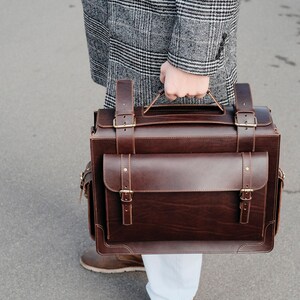 May include: A brown leather briefcase with brass buckles and a top handle. The briefcase is being carried by a person wearing a gray plaid coat and white pants.