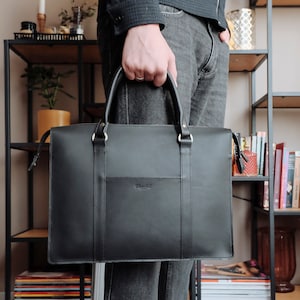 May include: A black leather briefcase with a gold zipper and a small pocket on the front. The briefcase has a handle and a strap.