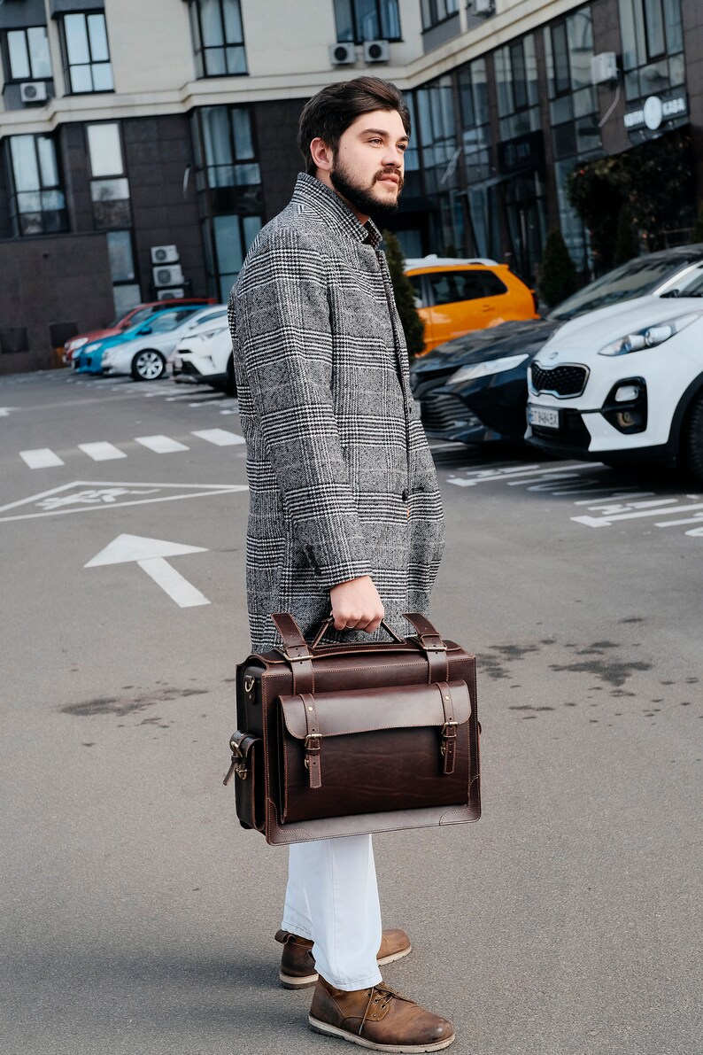 May include: A man in a gray plaid coat carries a brown leather briefcase with a top handle and multiple straps. He is walking on a city street.