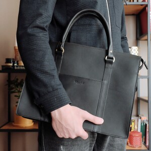 May include: A black leather briefcase with a top handle and a zippered closure. The briefcase has a subtle embossed logo on the front.
