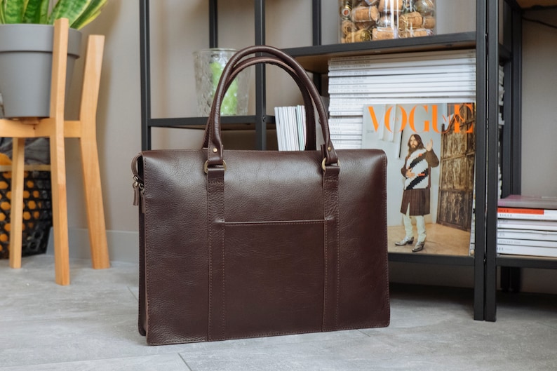 May include: A brown leather briefcase with a zipper and two handles. The briefcase is sitting on a wooden floor.