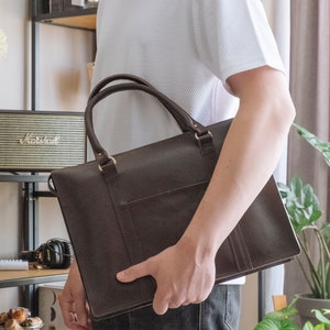May include: A brown leather briefcase with a top handle and a front pocket. The briefcase is held by a person wearing a white shirt and dark jeans.