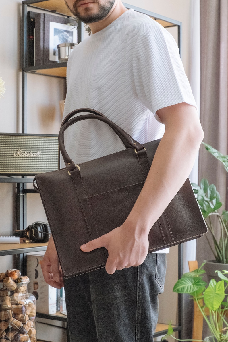 May include: A brown leather briefcase with a top handle and a front pocket. The briefcase is held by a person wearing a white shirt and dark jeans.