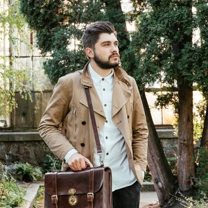 May include: A man wearing a beige jacket and a white button-down shirt is carrying a brown leather briefcase with a strap.