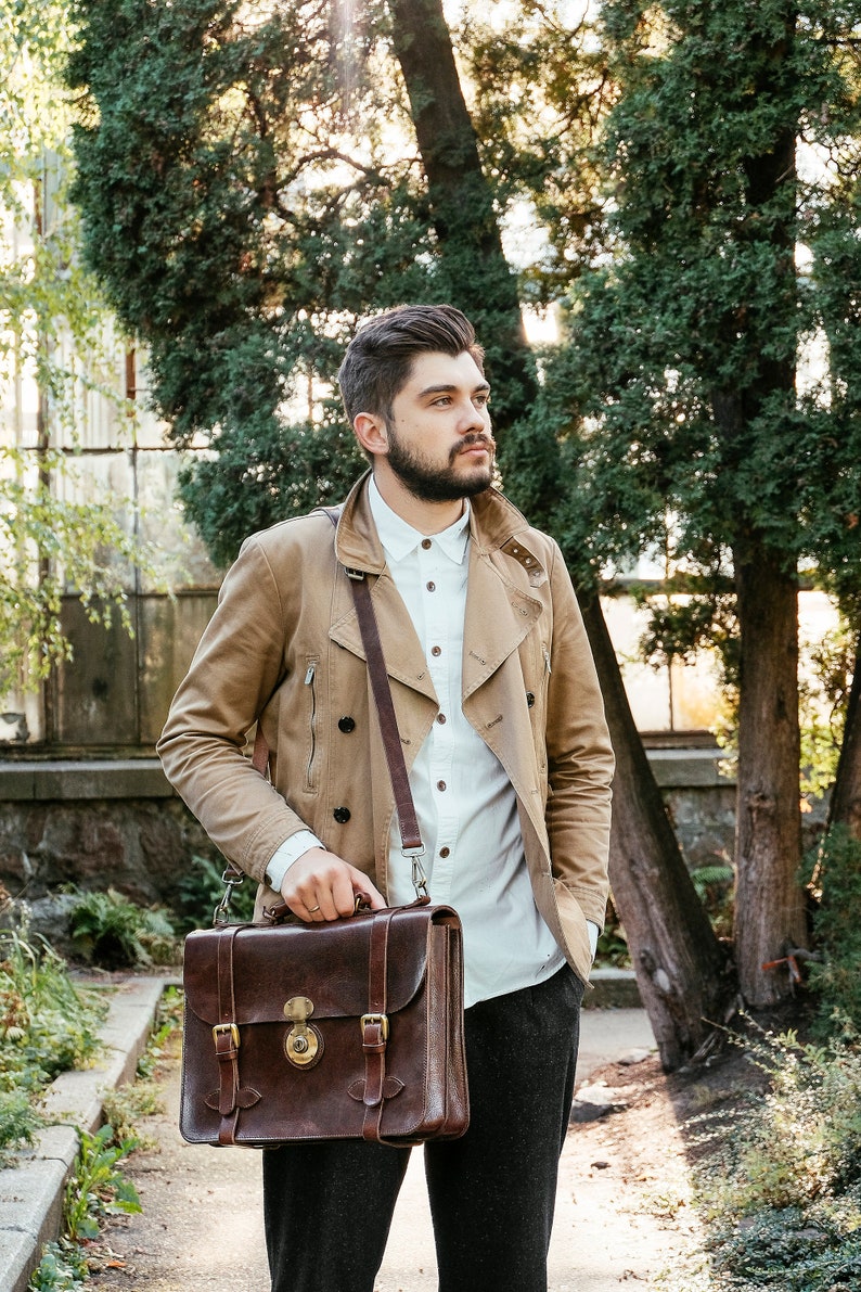 May include: A man wearing a beige jacket and a white button-down shirt is carrying a brown leather briefcase with a strap.