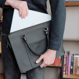 May include: A black leather briefcase with a zipper and a handle. A laptop computer is being placed inside the briefcase.