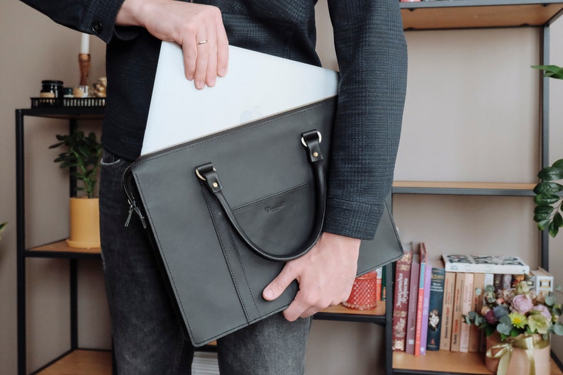 May include: A black leather briefcase with a zipper and a handle. A laptop computer is being placed inside the briefcase.