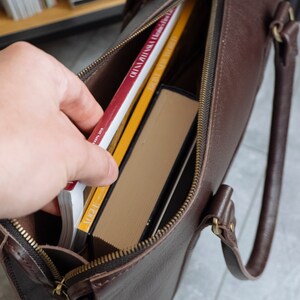 May include: A brown leather tote bag with a zipper closure. The bag is open and filled with books. The books have red, yellow, and white covers.