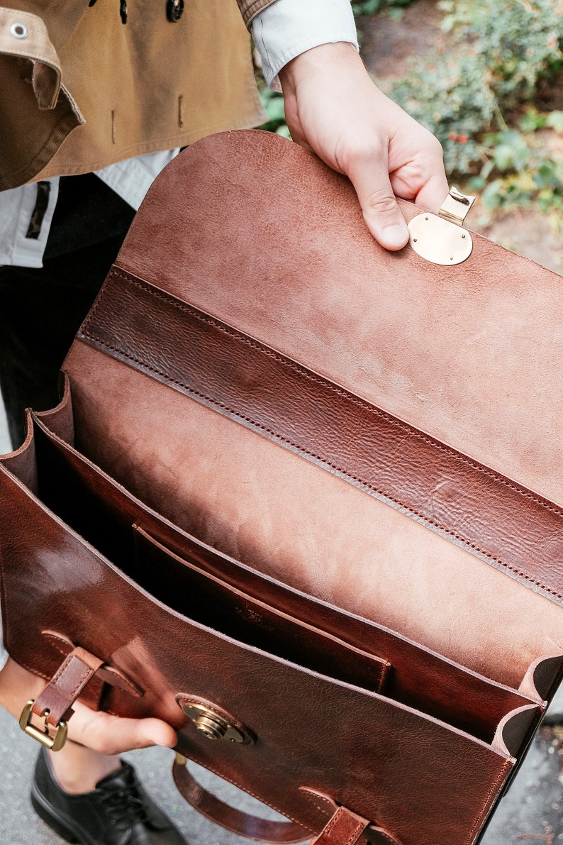 May include: A brown leather briefcase with a brass clasp and buckle. The briefcase is open and the inside is lined with brown leather.