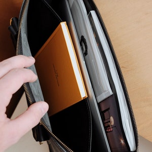 May include: A black leather briefcase with a zipper closure. The briefcase is open and contains a yellow notebook with the text "A.P.M.  FAMILY & COLLECTION" and a stack of papers.