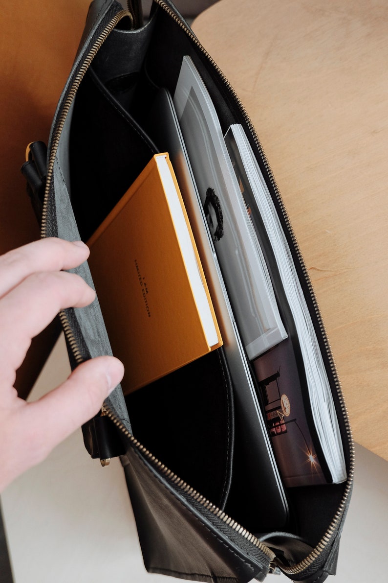May include: A black leather briefcase with a zipper closure. The briefcase is open and contains a yellow notebook with the text "A.P.M.  FAMILY & COLLECTION" and a stack of papers.