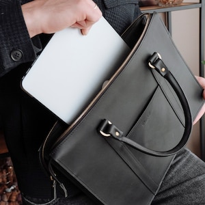 May include: A black leather briefcase with a gold zipper and a gold buckle. A laptop computer is being placed inside the briefcase.