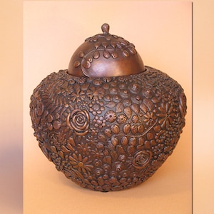 May include: A brown, handcrafted ceramic jar with a lid. The jar is covered in a detailed floral pattern, including roses, daisies, and leaves. The lid has a small knob and a decorative floral border. The jar is a decorative piece.
