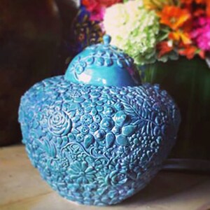 K&ouml;nnte beinhalten: Eine tealblaue Keramikvase mit floralem Muster. Die Vase ist mit einem erhabenen Blumenmuster verziert und hat eine abgerundete Form.