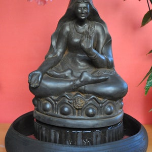 May include: Black stone statue of a seated Buddha figure with a crown and a hand gesture. The statue is placed on a round black base with a decorative rim. The base is sitting on a larger black bowl with a decorative rim.