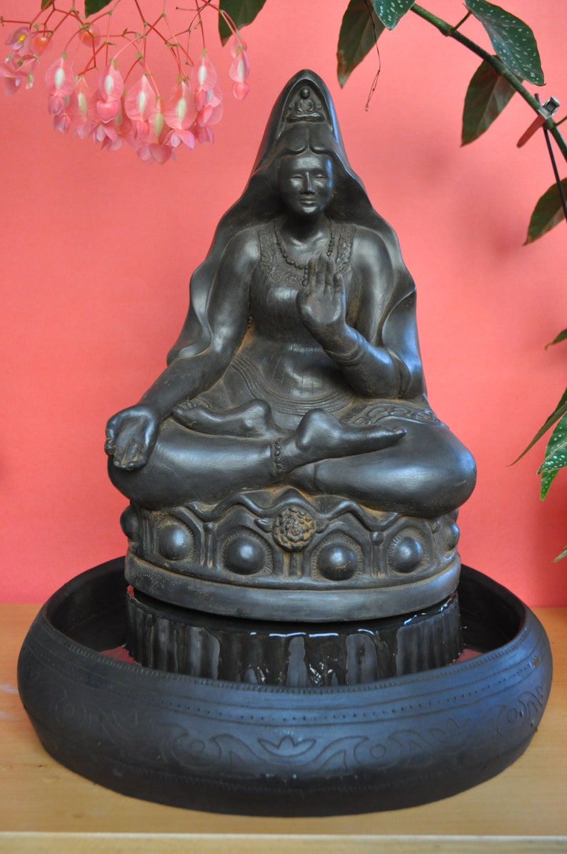 May include: Black stone statue of a seated Buddha figure with a crown and a hand gesture. The statue is placed on a round black base with a decorative rim. The base is sitting on a larger black bowl with a decorative rim.
