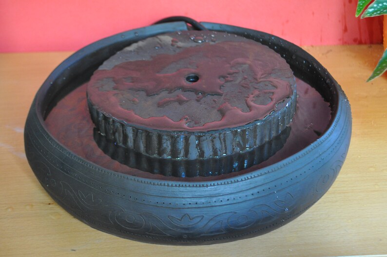 May include: A black ceramic fountain with a round, textured top. The fountain is filled with water and has a small hole in the centre of the top.