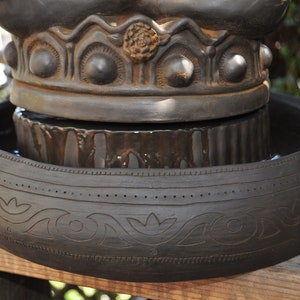May include: A black ceramic fountain with a decorative top and a lower bowl with intricate carvings. The fountain is filled with water and has a rustic look.