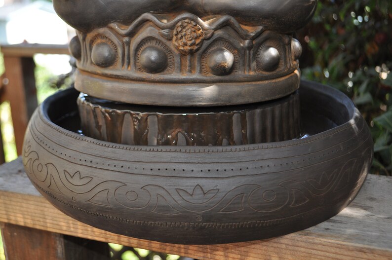 May include: A black ceramic fountain with a decorative top and a lower bowl with intricate carvings. The fountain is filled with water and has a rustic look.