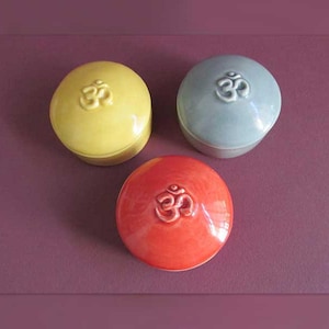 May include: Three small ceramic containers with lids. Each container is a different color: yellow, gray, and red. The lids have a raised "Om" symbol.