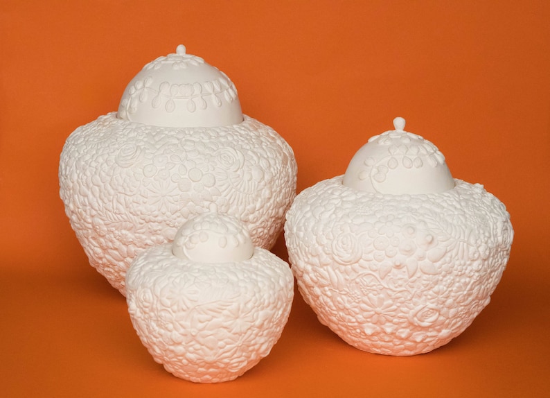 K&ouml;nnte beinhalten: Drei wei&szlig;e Keramikurnen mit aufwendigen Blumendesigns. Die Urnen sind in verschiedenen Gr&ouml;&szlig;en und in einer Reihe auf einem einfarbigen orangefarbenen Hintergrund angeordnet.