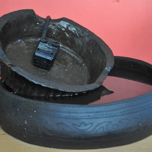 May include: A black ceramic fountain with a small pump and a decorative design. The fountain is filled with water and has a rustic look.