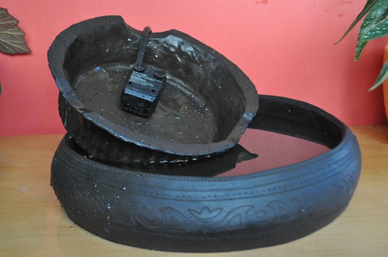 May include: A black ceramic fountain with a small pump and a decorative design. The fountain is filled with water and has a rustic look.
