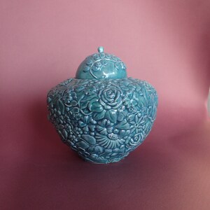K&ouml;nnte beinhalten: Eine tealfarbene Keramikvase mit Deckel, aufwendig verziert mit erhabenen Blumendesigns. Die Vase hat eine abgerundete Form und eine glatte, gl&auml;nzende Oberfl&auml;che.