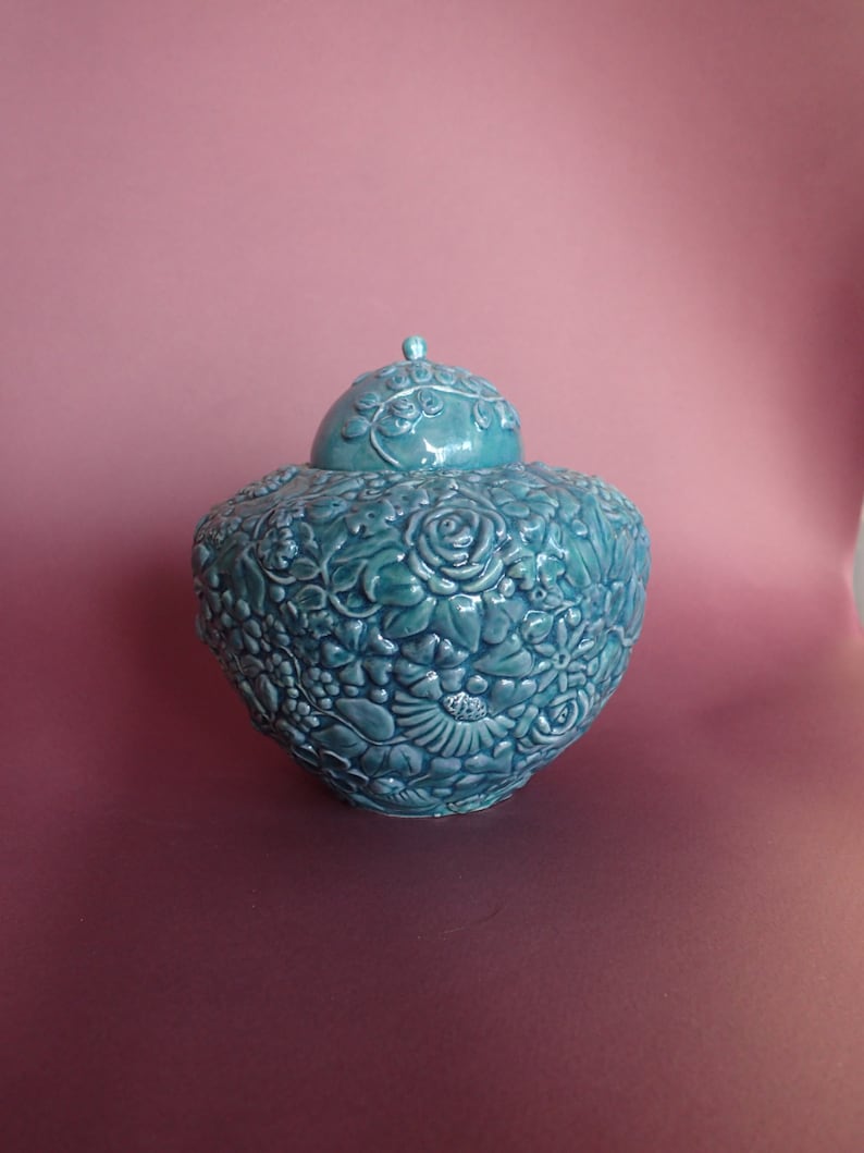 K&ouml;nnte beinhalten: Eine tealfarbene Keramikvase mit Deckel, aufwendig verziert mit erhabenen Blumendesigns. Die Vase hat eine abgerundete Form und eine glatte, gl&auml;nzende Oberfl&auml;che.