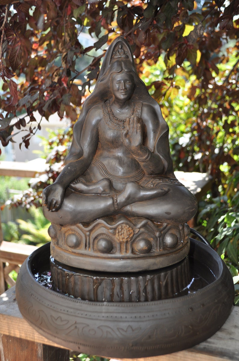 May include: A dark brown ceramic statue of a seated Buddha with a flowing robe, placed on a tiered fountain base with water flowing over the tiers. The statue has a serene expression and is adorned with intricate details.