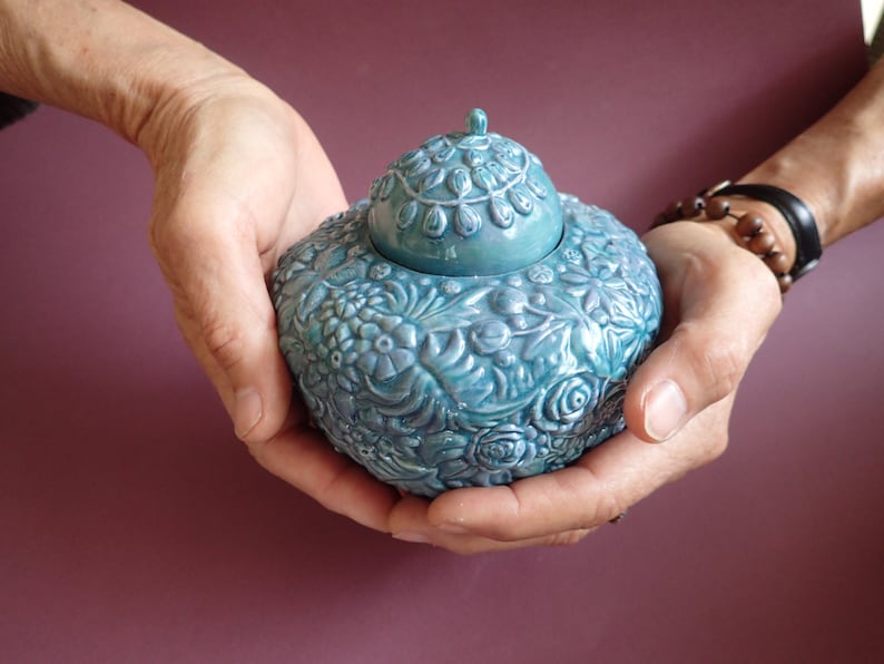 K&ouml;nnte beinhalten: Eine tealblaue Keramikdose mit floralem Muster und Deckel. Die Dose wird in zwei H&auml;nden gehalten.