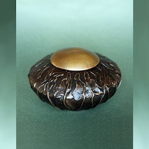 Puede incluir: Un recipiente decorativo artesanal de color bronce con una tapa dorada. El recipiente tiene un diseño texturizado y orgánico. La tapa tiene una forma lisa y redondeada. El recipiente está sobre una superficie verde.