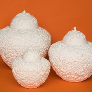 May include: Three white ceramic urns with intricate floral designs. The urns are of varying sizes and are arranged in a row on a solid orange background.