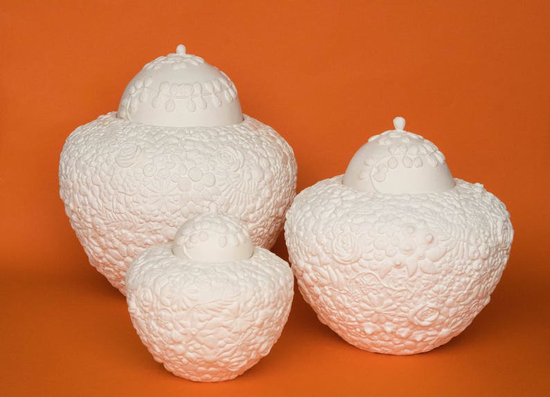 May include: Three white ceramic urns with intricate floral designs. The urns are of varying sizes and are arranged in a row on a solid orange background.