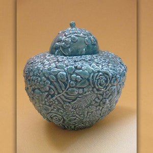 May include: A teal ceramic jar with a lid, decorated with a floral design. The jar is intricately detailed with raised flowers and leaves.
