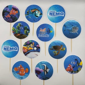 Peut inclure: Une collection de décorations de cupcakes sur le thème du Monde de Nemo. Chaque décoration ronde présente des personnages comme Nemo, Dory et Crush, sur un fond bleu océan. Certaines décorations portent l'inscription "Finding Nemo" ou "Just Keep Swimming".