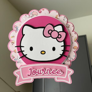 May include: A pink and white Hello Kitty-themed decoration with the word "Journee" on a banner. The design features a scalloped edge and a layered effect, with a clear plastic stick for display.