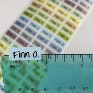 May include: A sheet of small, rectangular adhesive labels in various pastel colors, each printed with the name Finn O. in black text. A clear plastic ruler is positioned over one label, showing it measures approximately 0.5 inches in width.