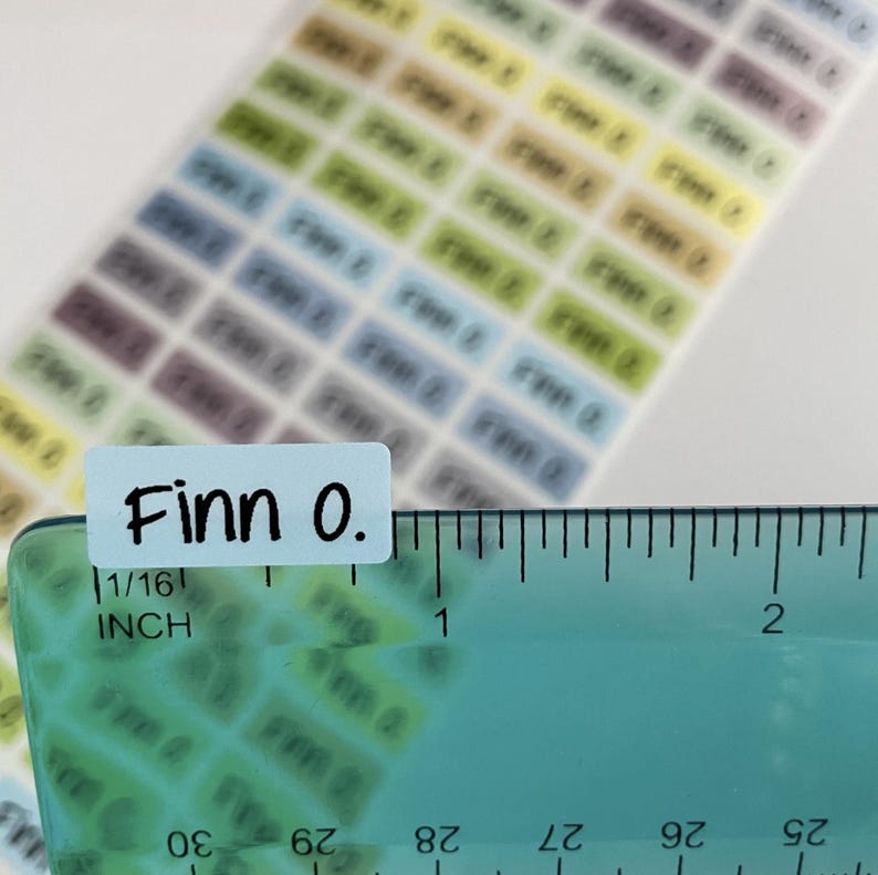 May include: A sheet of small, rectangular adhesive labels in various pastel colors, each printed with the name Finn O. in black text. A clear plastic ruler is positioned over one label, showing it measures approximately 0.5 inches in width.